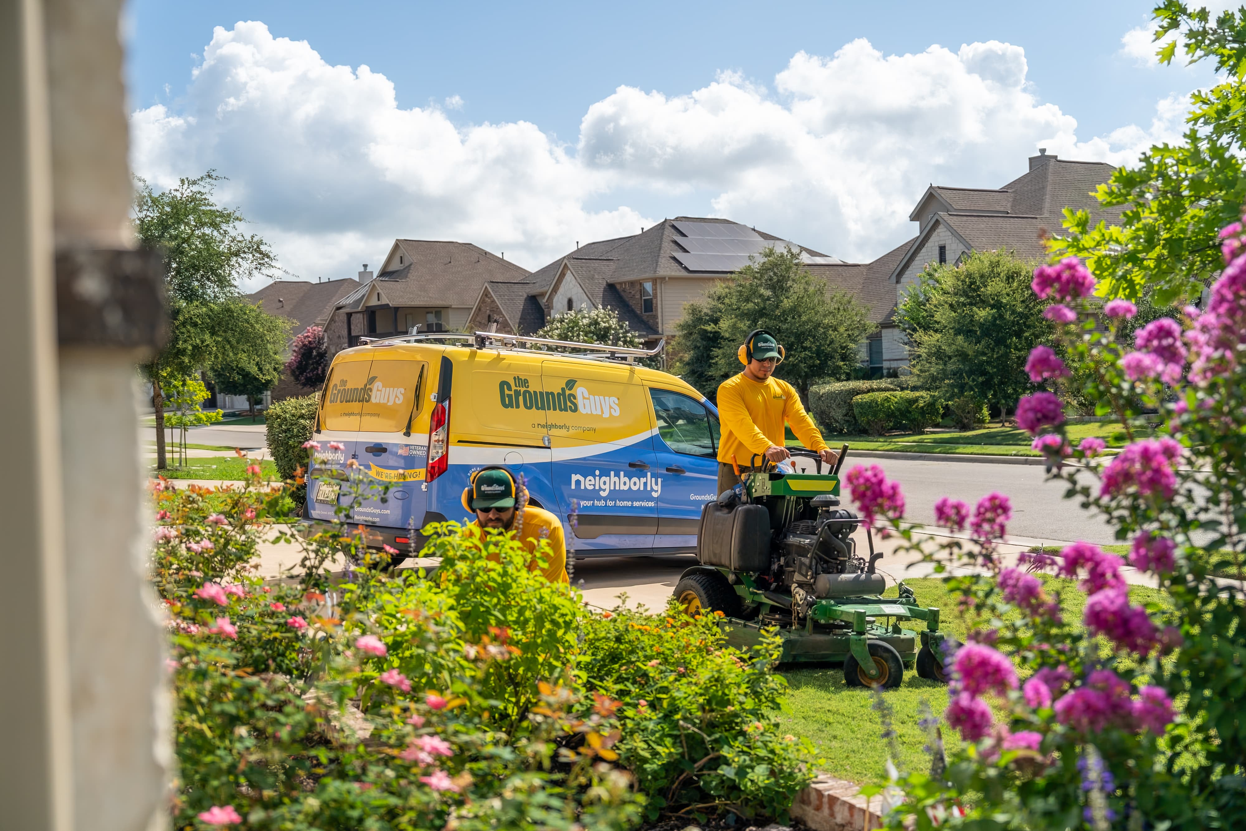 Yellow and blue "The Grounds Guys" van with "Neighborly" livery, parked next to a man mowing lawn.