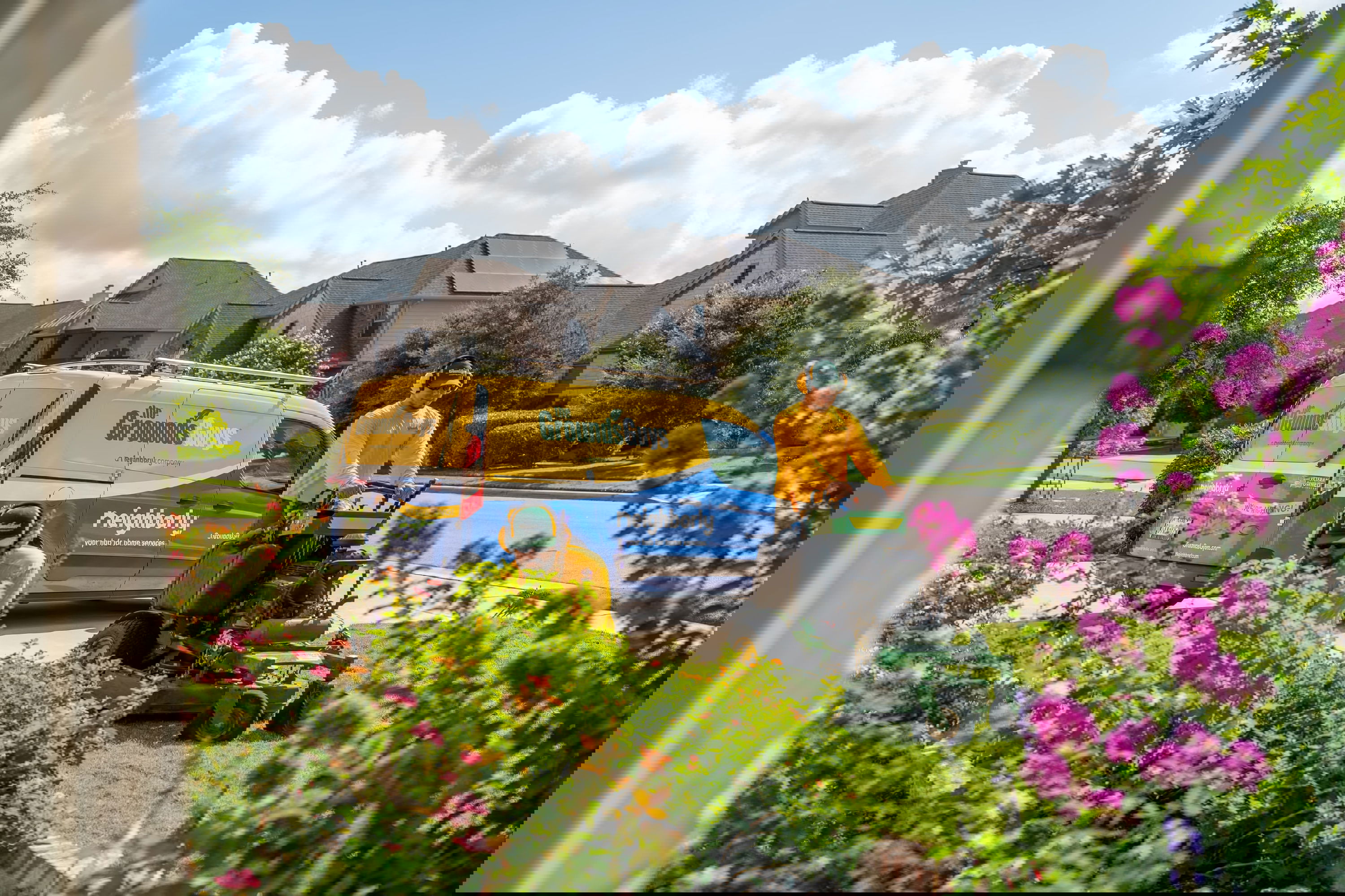 Yellow and blue "The Grounds Guys" van with "Neighborly" livery, parked next to a man mowing lawn.