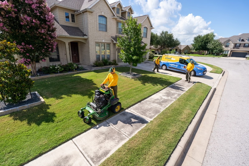 Grounds Guys commercial van in blue with yellow livery, parked by a manicured lawn and house.