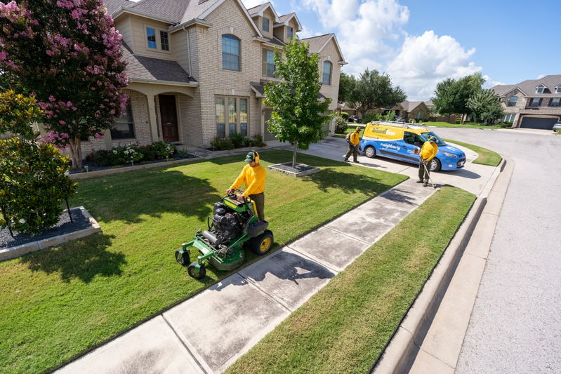 Grounds Guys commercial van in blue with yellow livery, parked by a manicured lawn and house.