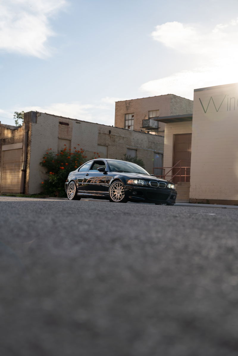 Black 2002 BMW M3 coupe on gold multi-spoke rims parked near buildings.