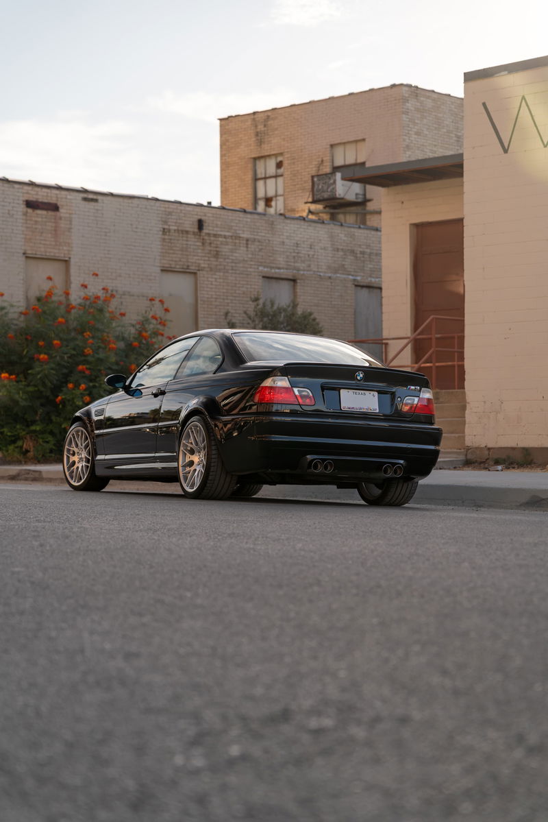 Black 2002 BMW M3 E46 coupe with aftermarket multi-spoke rims and quad exhaust tips.