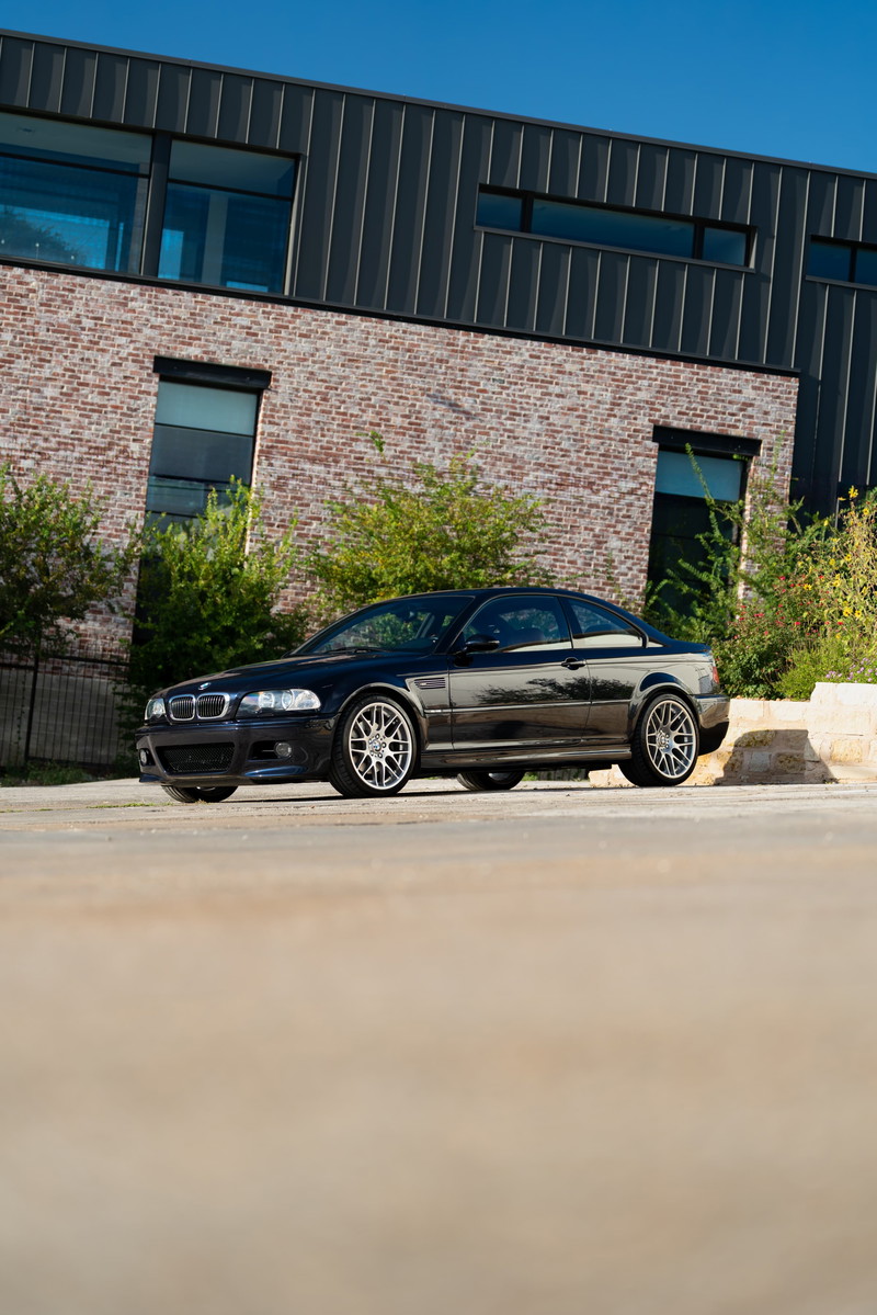 Black 2002 BMW M3 coupe with silver rims parked in front of a modern building.