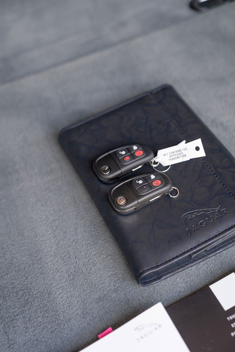 Two Jaguar XJR key fobs rest on a navy leather owner's manual. Radiance Red 2006 XJR.