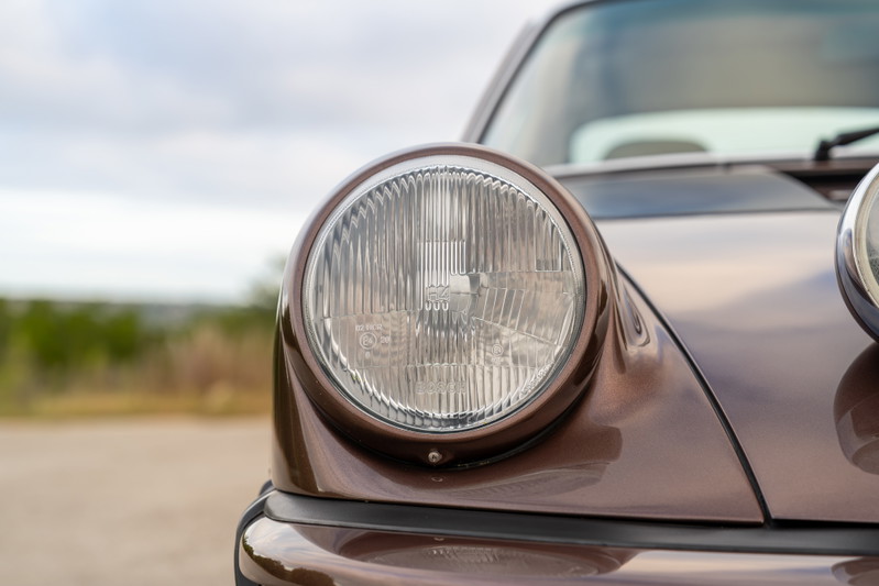 Close-up of the iconic round headlight on a copper brown metallic 1975 Porsche 911 Carrera.