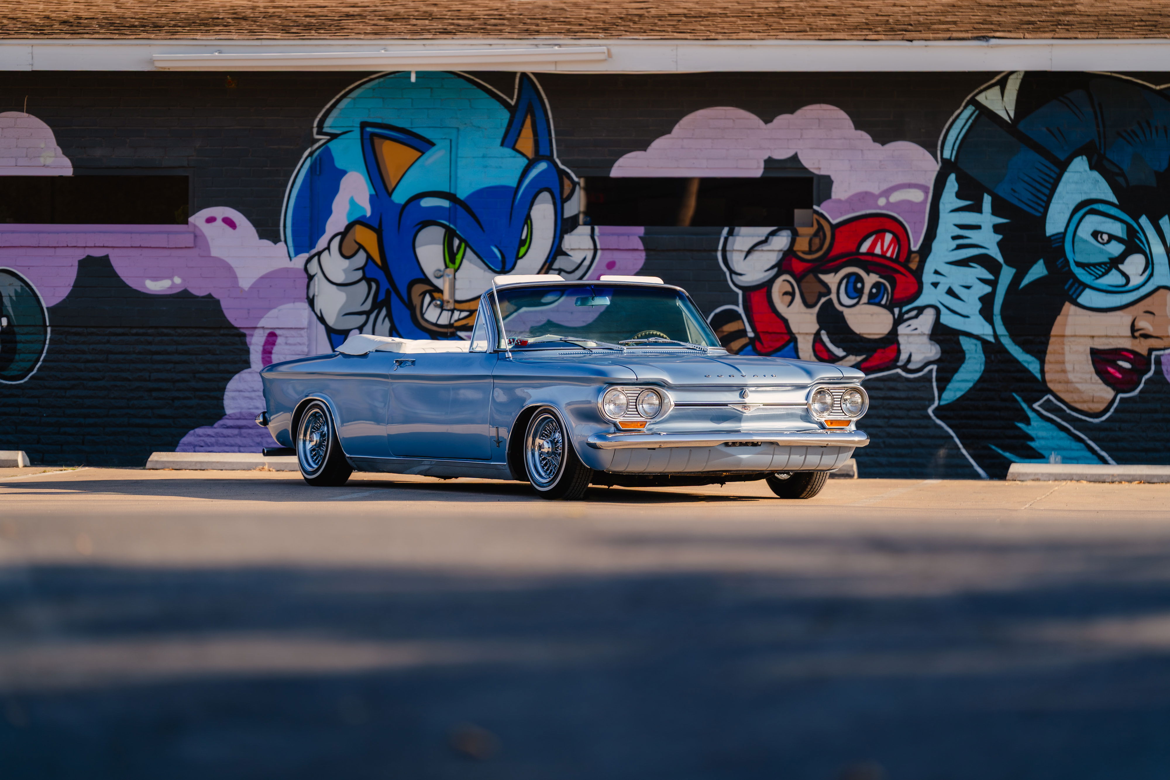 1966 Chevrolet Corvair convertible in blue metallic with chrome rims, parked in front of a mural.
