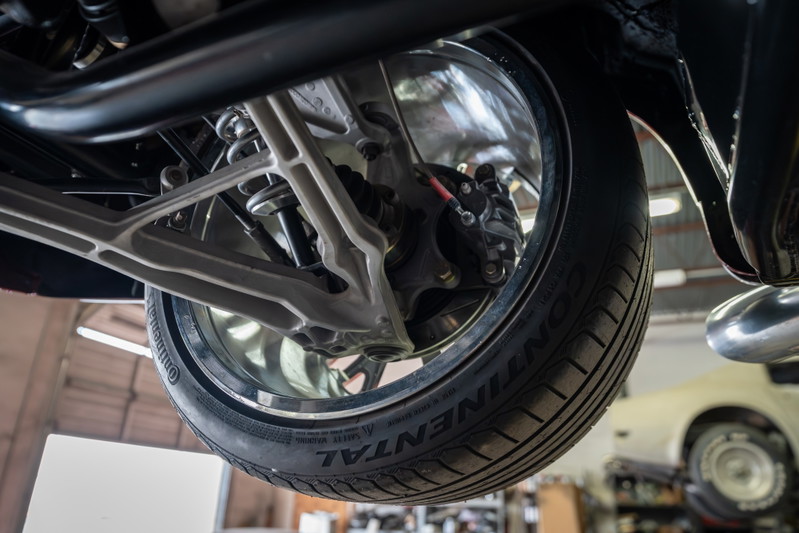 Close-up of a 1965 Chevrolet Corvette's rear suspension, showing alloy control arms, coilover shock, and Continental tire on polished rim.