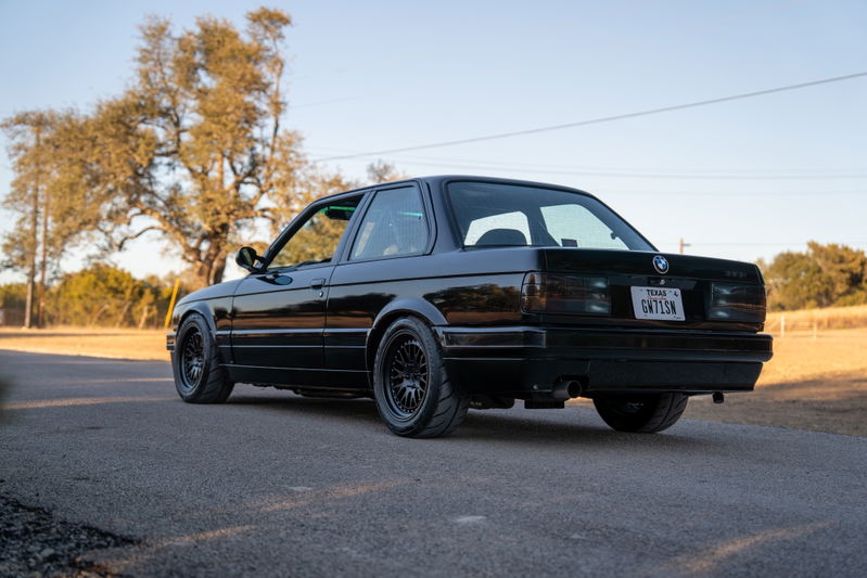 Rear view of a black 1989 BMW 325is with black mesh rims and tinted taillights.