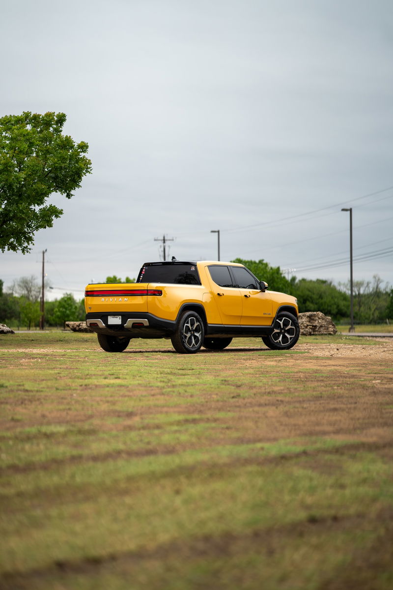Yellow 2022 Rivian R1T pickup truck with black trim and sport rims parked on grass.