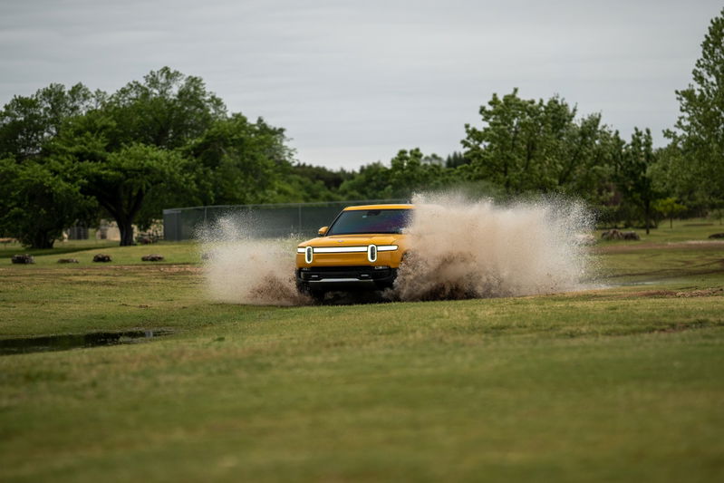 Yellow Rivian R1T SUV kicking up mud and water off-road.