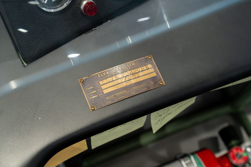 Close-up of a brass Elva Porsche 4-Cam chassis plate on a gray dashboard.