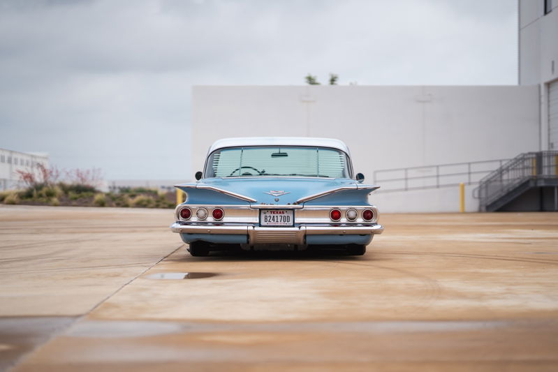 Rear view of a light blue 1960 Chevrolet Impala lowrider with prominent tail fins and chrome bumper.
