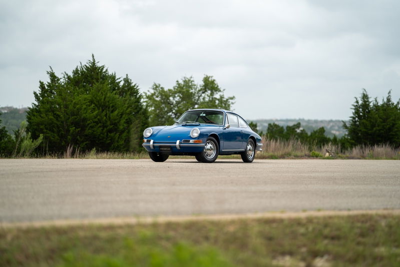 A blue 1965 Porsche 912 coupe sits parked on an asphalt road, with chrome rims and trim.