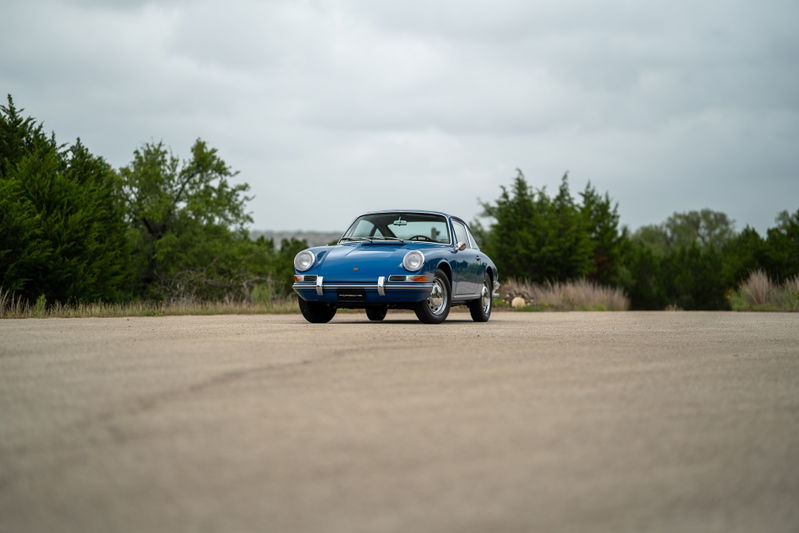 Blue 1965 Porsche 912 coupe, front view, on paved lot with trees.