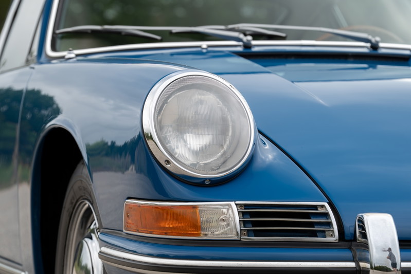 Close up of a blue 1965 Porsche 912 fender, showing chrome trim, headlight, and amber turn signal.