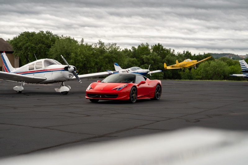 A red Ferrari 458 Italia with black rims parked on an asphalt runway next to a small airplane.